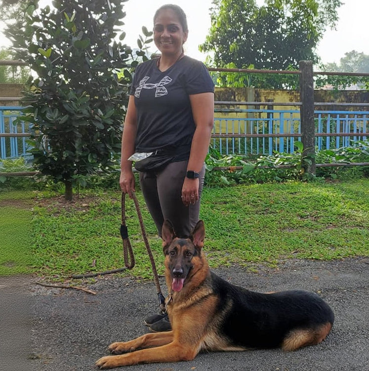 Woman standing next to a German Shepherd dog on a leash in an outdoor setting.